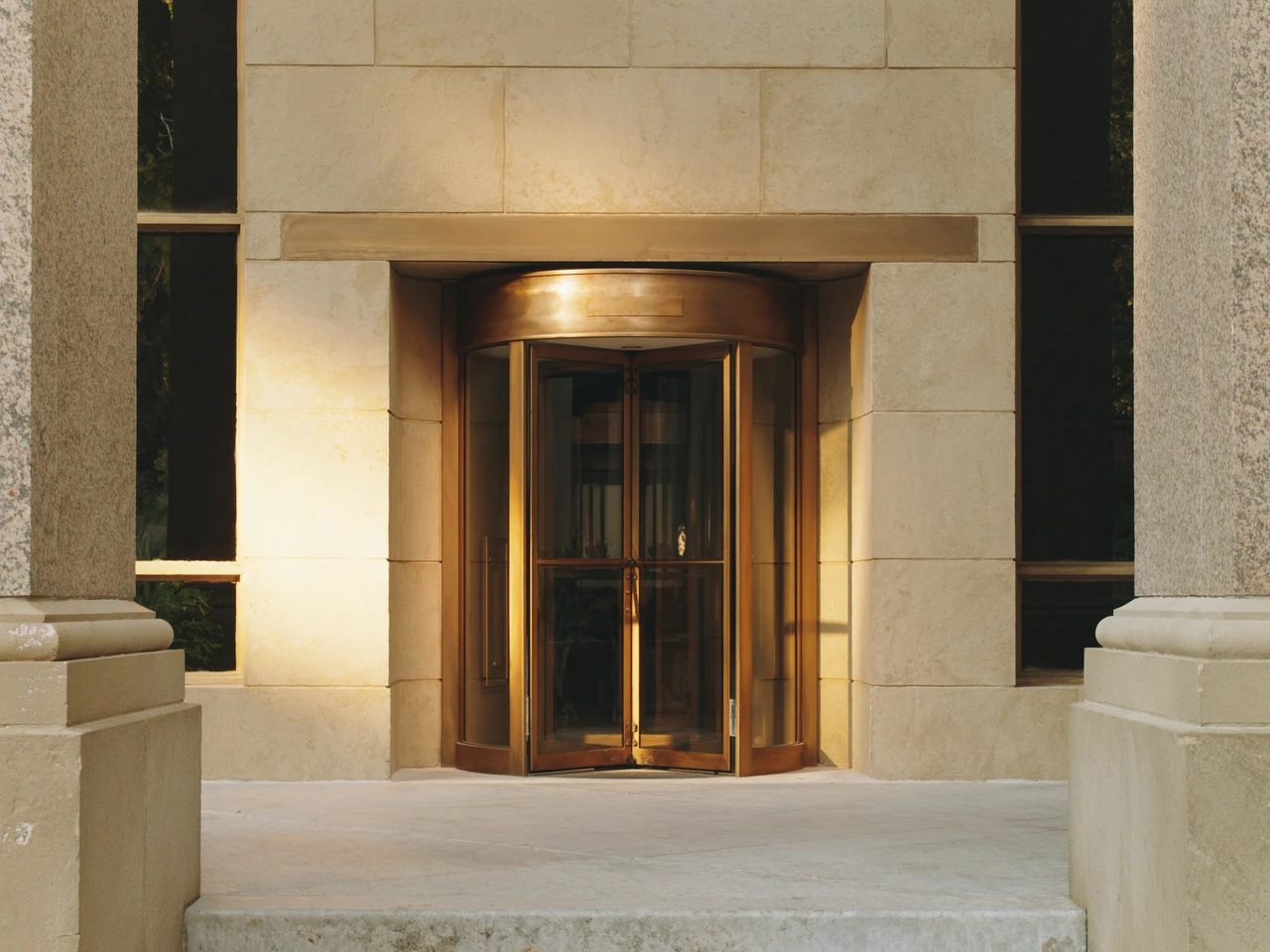 Illustrative stone bank entrance with a bronze revolving door in afternoon sunlight.